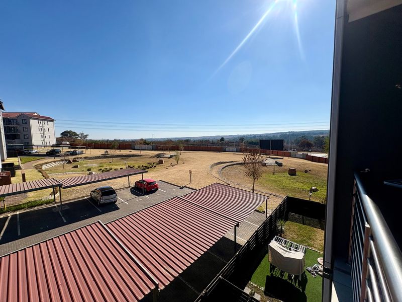 Elevated view overlooking the apartment complex grounds, featuring covered parking bays, landscaped open spaces, and perimeter fencing under a clear sky.