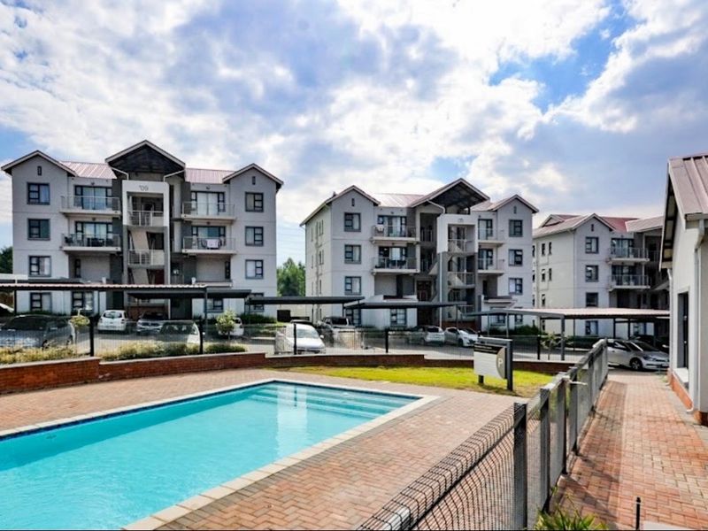A well-maintained communal swimming pool area featuring brick paving, metal fencing, and views of the apartment complex with covered parking bays.