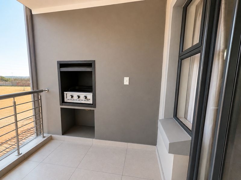 A modern, covered balcony featuring tiled flooring, a built-in braai, and a stainless steel balustrade with views of the surrounding area.