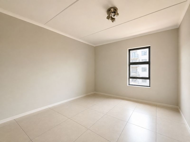 A bright, empty bedroom featuring neutral wall tones, large format floor tiles, and a modern ceiling-mounted spotlight fixture.