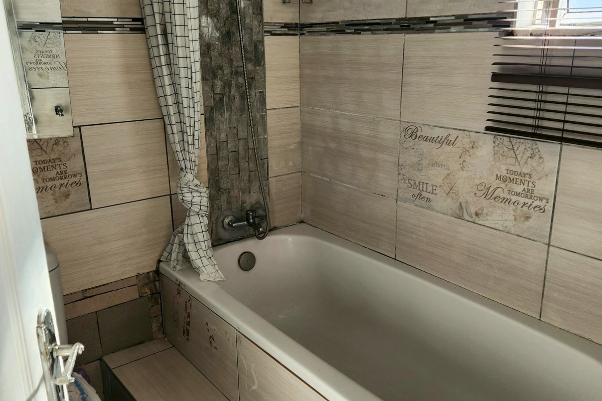 Compact bathroom featuring a built-in bathtub with shower attachment, tiled walls, and a window with venetian blinds. Requires maintenance and cosmetic updates.