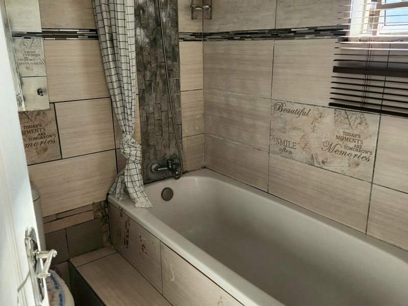 Compact bathroom featuring a built-in bathtub with shower attachment, tiled walls, and a window with venetian blinds. Requires maintenance and cosmetic updates.