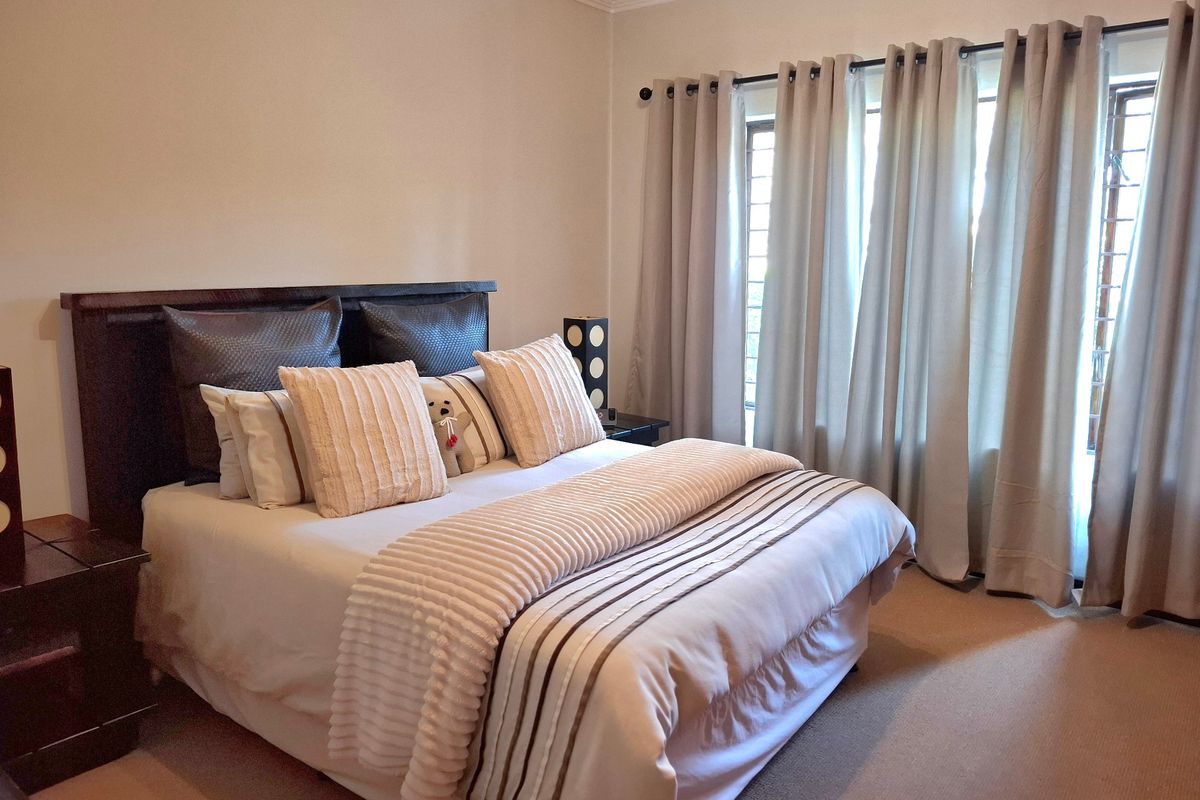 A well-maintained bedroom featuring neutral wall tones, carpeted flooring, and large windows dressed with full-length curtains.