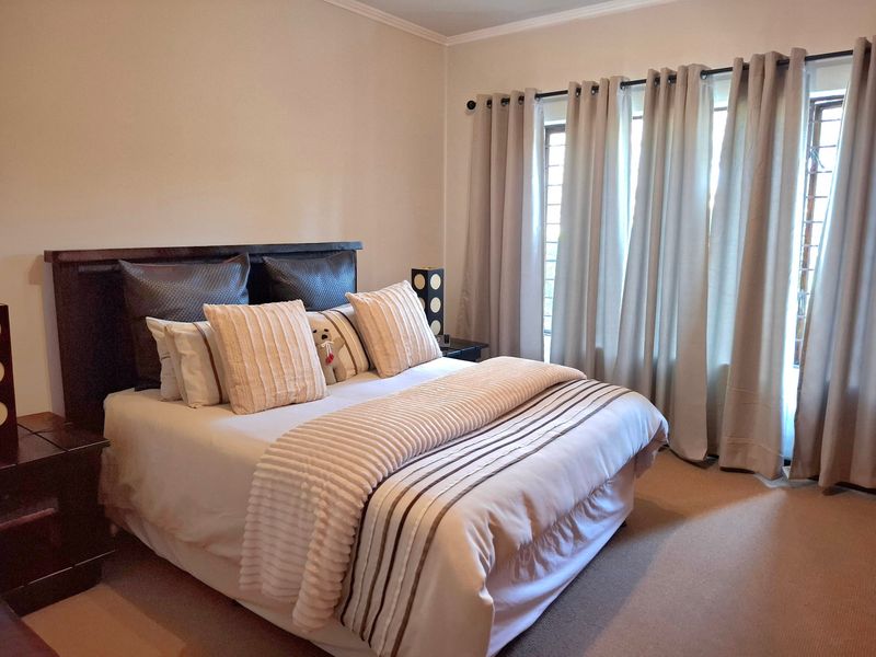 A well-maintained bedroom featuring neutral wall tones, carpeted flooring, and large windows dressed with full-length curtains.