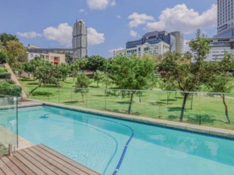 A well-maintained swimming pool featuring a modern glass balustrade and timber decking, overlooking a landscaped park area with city skyline views.