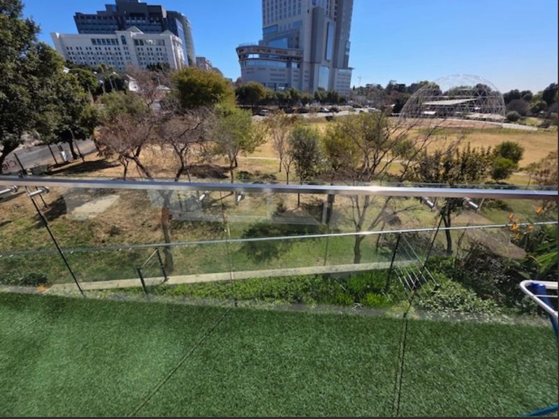 A balcony featuring a modern glass balustrade with a stainless steel handrail, overlooking a park and urban cityscape.