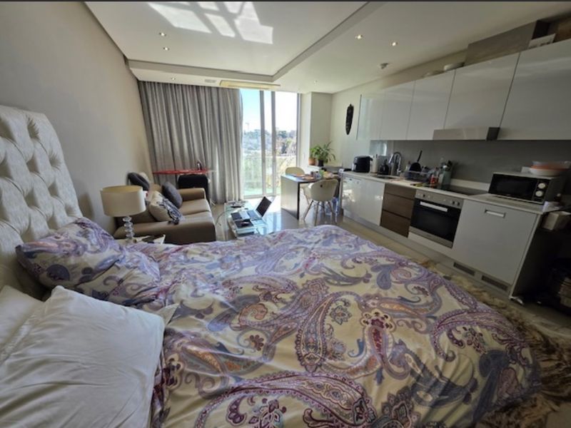 A modern open-plan studio apartment featuring a compact kitchenette with built-in appliances and direct access to a balcony through sliding glass doors.