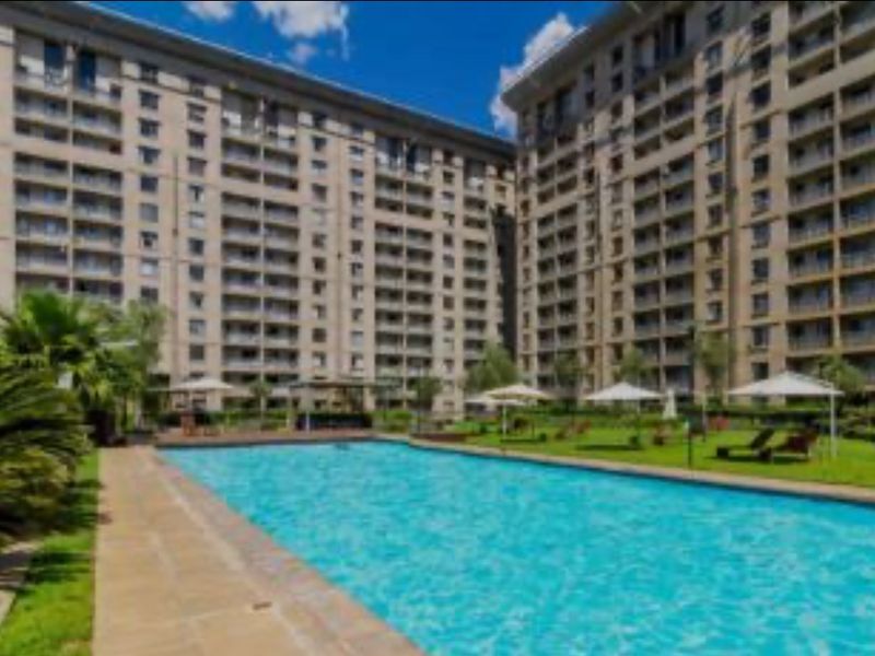 A well-maintained communal swimming pool area surrounded by landscaped gardens and the facade of a modern multi-story apartment complex.