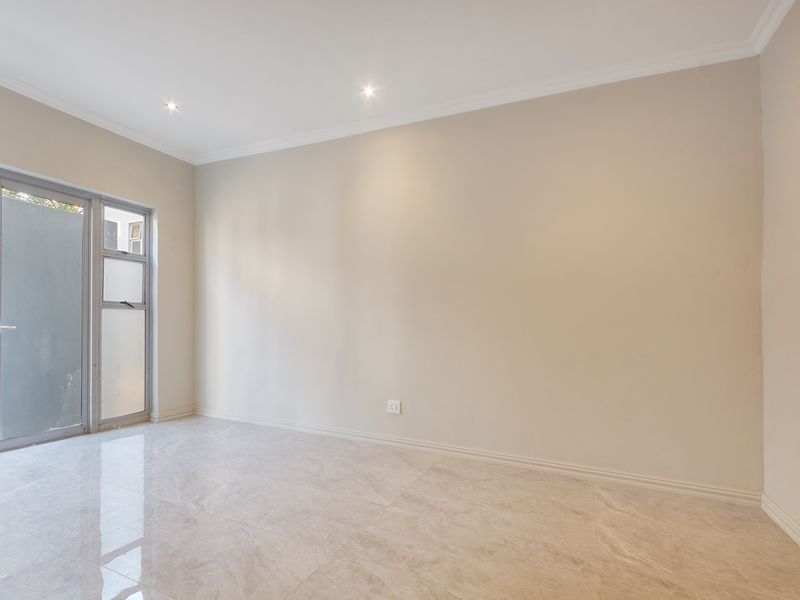 A bright, empty living area featuring neutral tiled flooring, recessed downlighting, and a glass door providing access to the exterior.