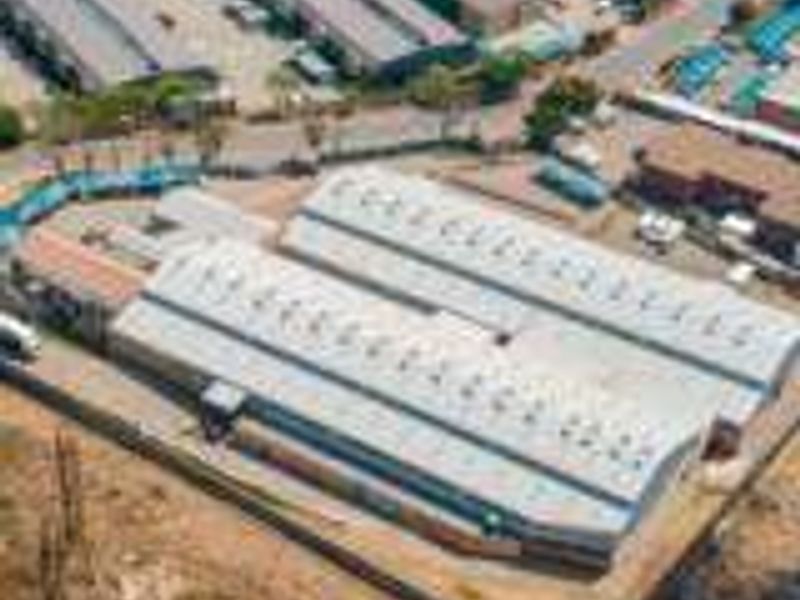 Aerial view of an industrial warehouse property featuring a large metal roof, perimeter fencing, and paved yard space within an industrial park setting.