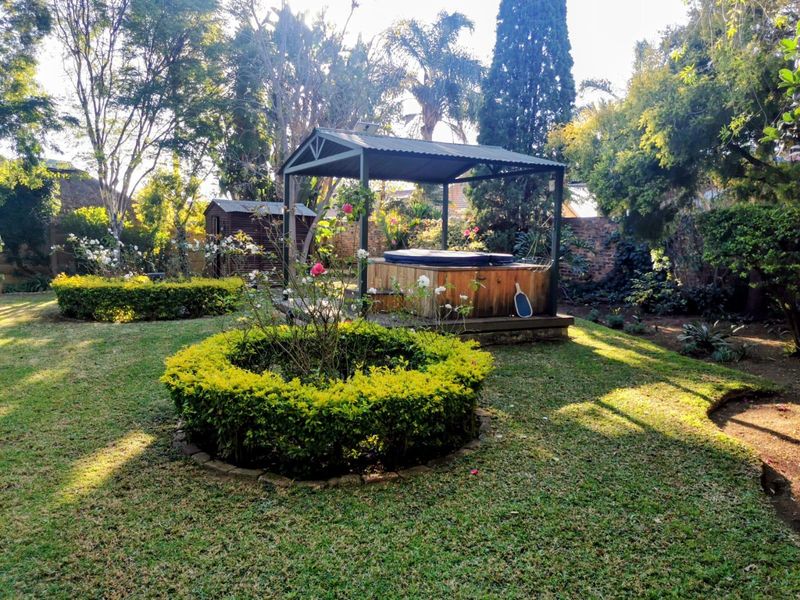 A well-maintained garden featuring a landscaped lawn, mature trees, a covered gazebo housing a hot tub, and a small garden shed.
