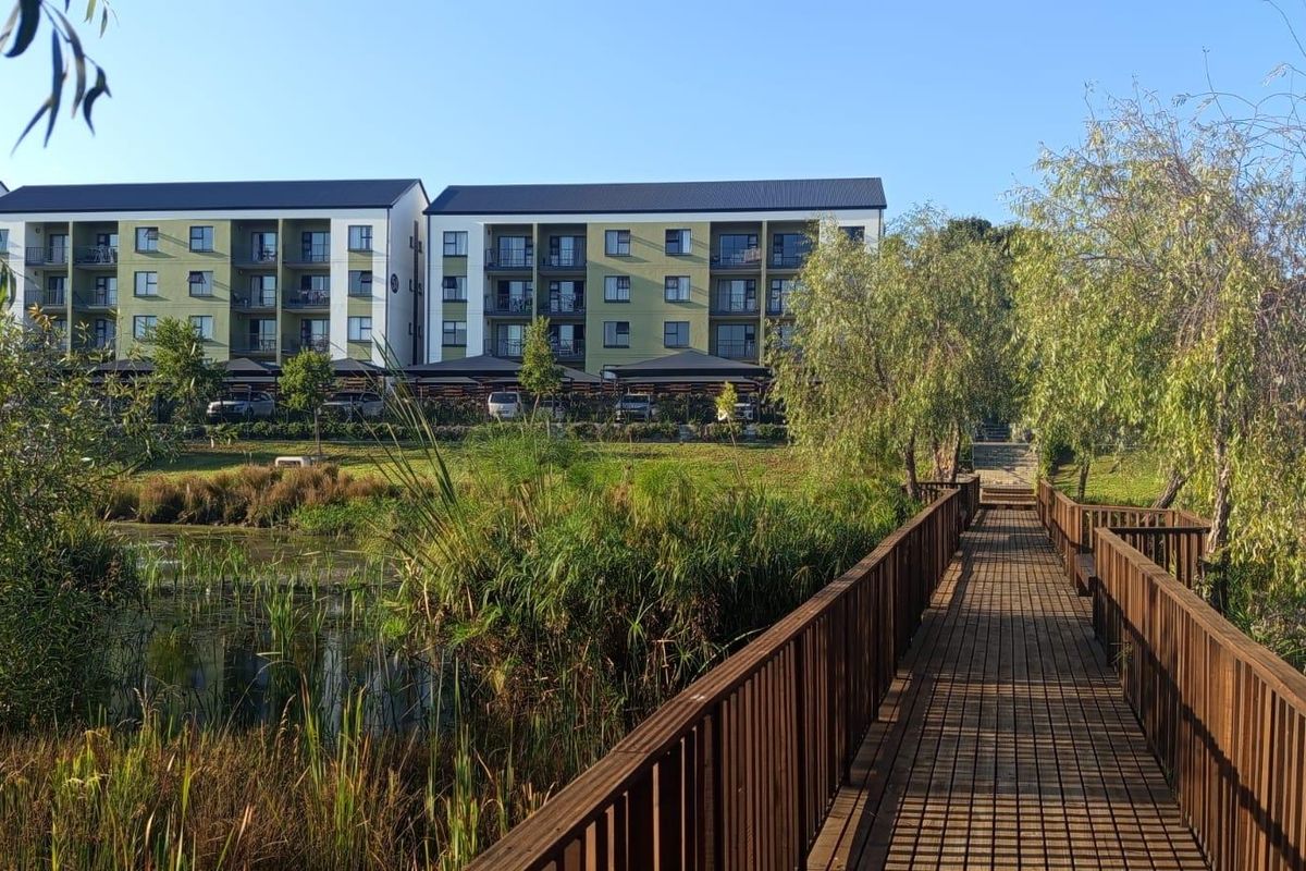 A scenic view of a multi-story apartment complex featuring a wooden boardwalk leading through a landscaped wetland area.