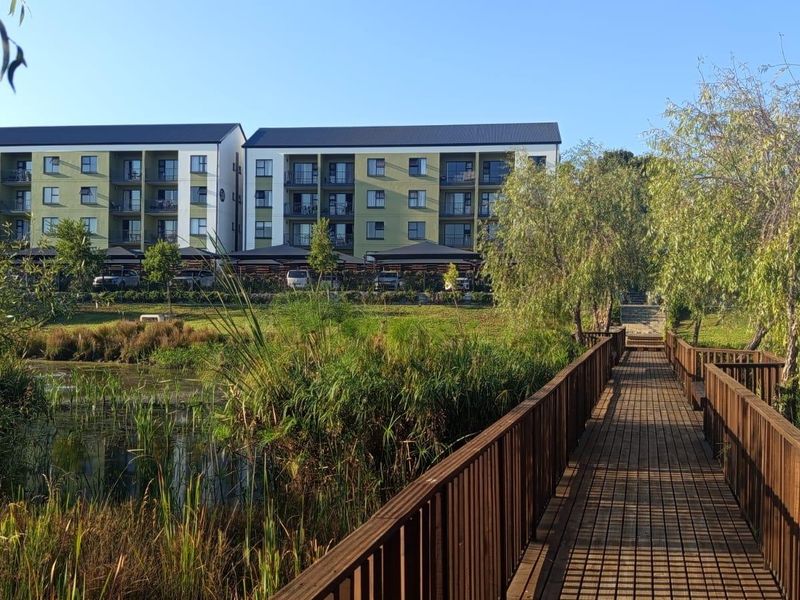 A scenic view of a multi-story apartment complex featuring a wooden boardwalk leading through a landscaped wetland area.