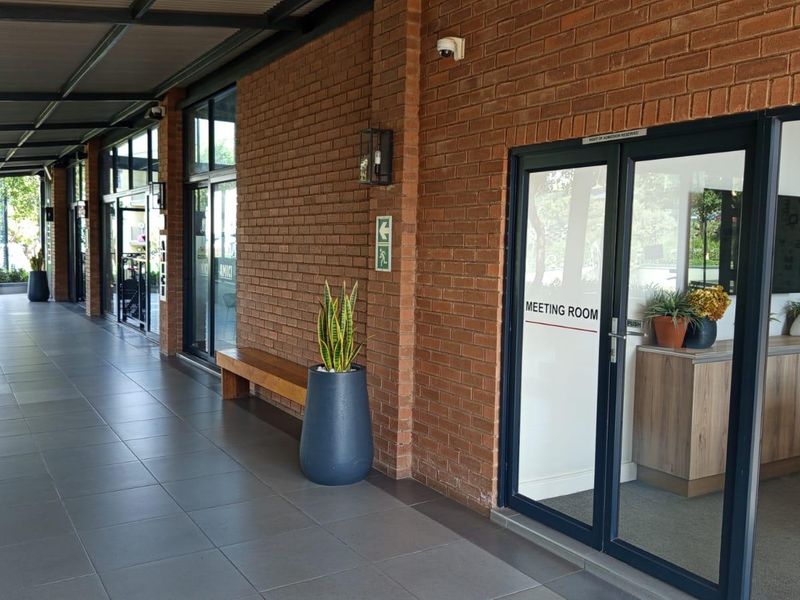 A covered outdoor walkway featuring exposed brick walls, tiled flooring, and glass-paneled access doors leading to a meeting room.