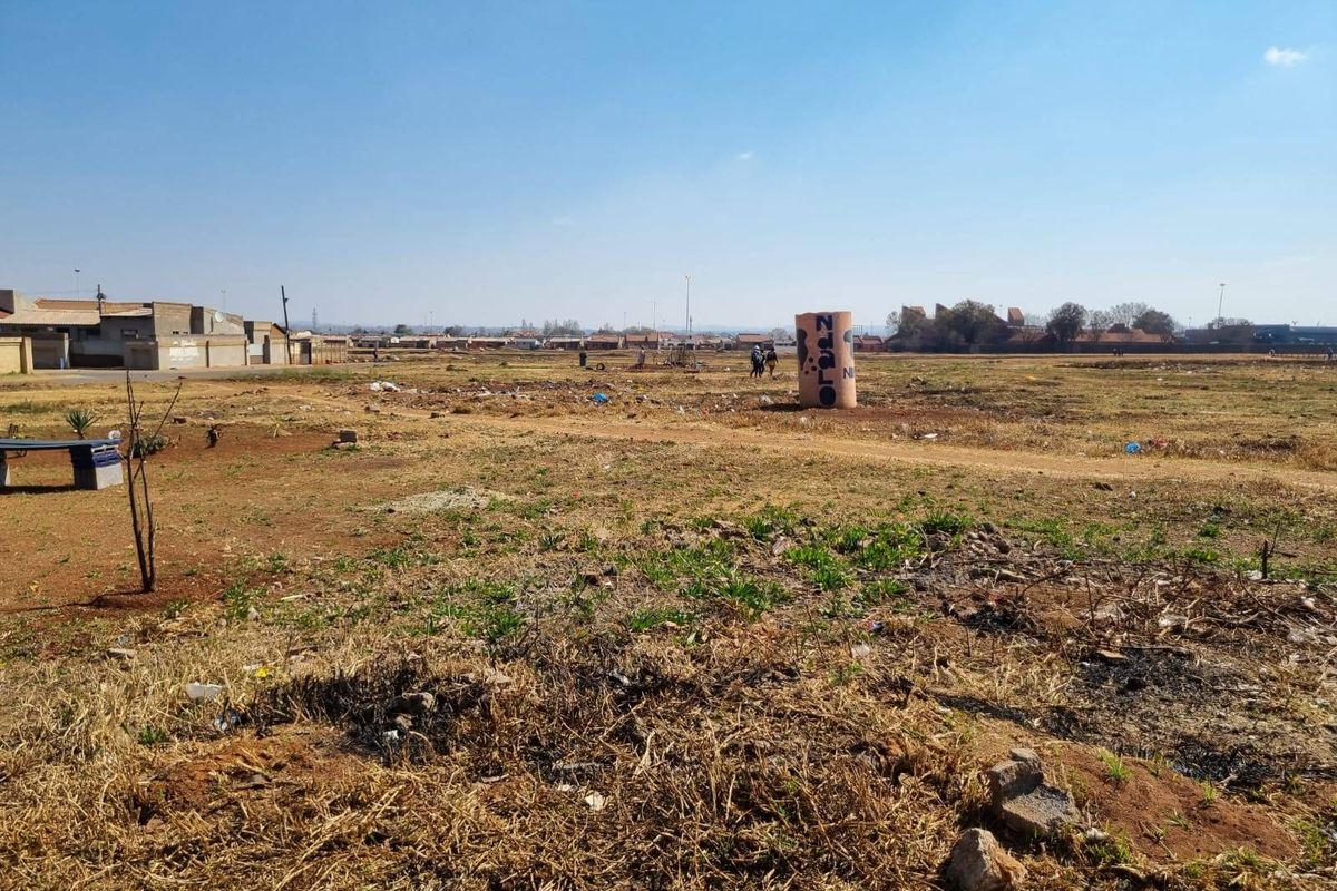 A wide-angle view of a vacant residential plot of land with sparse vegetation and surrounding neighborhood infrastructure.