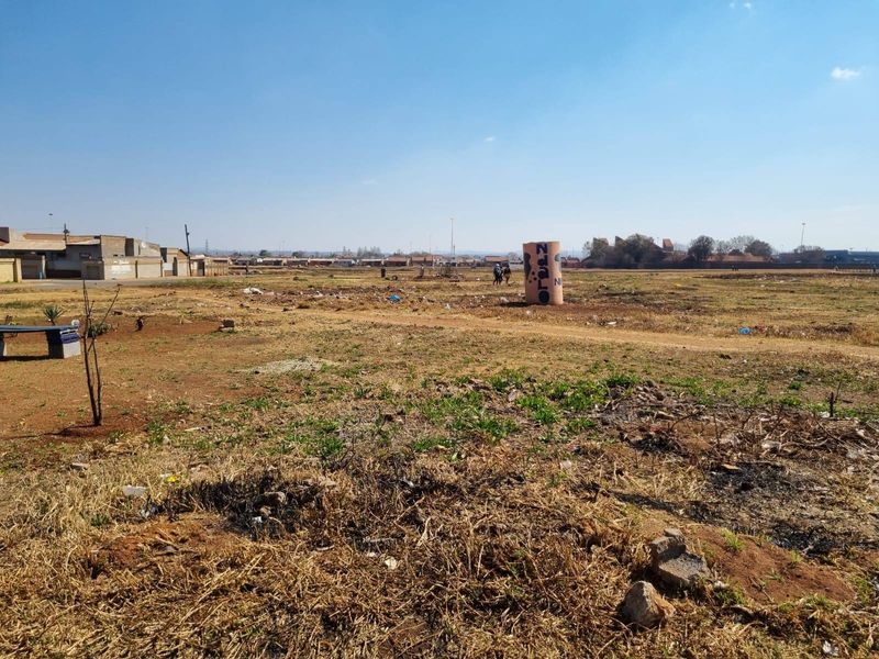 A wide-angle view of a vacant residential plot of land with sparse vegetation and surrounding neighborhood infrastructure.