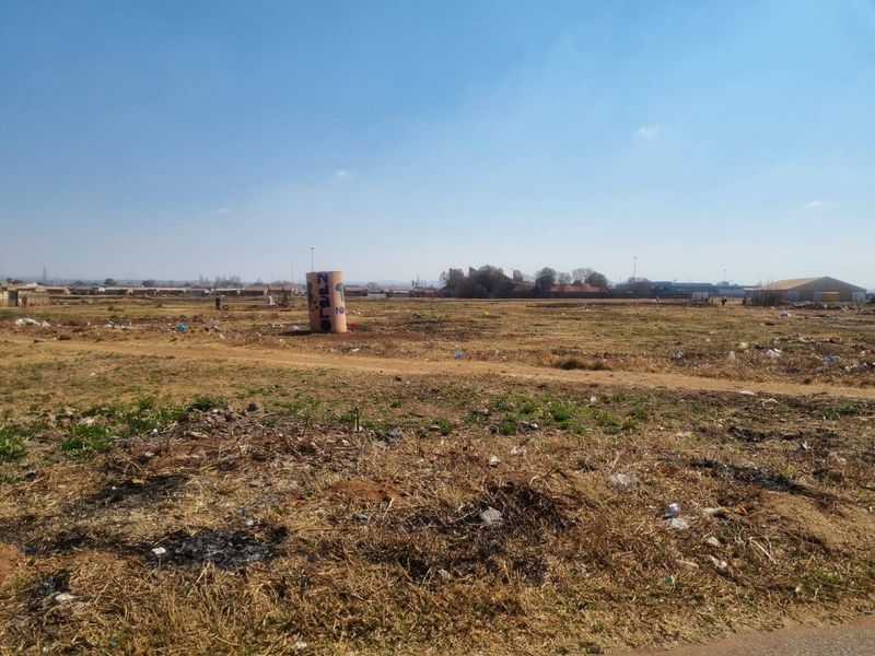 A wide-angle view of a vacant residential plot featuring dry grass and a small utility structure in the distance.