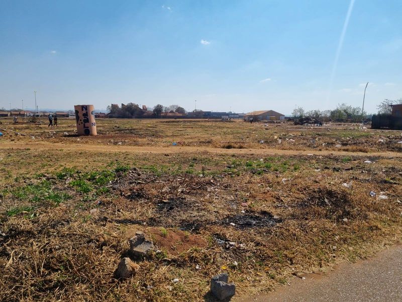 A wide-angle view of a vacant residential plot featuring dry, unmaintained ground and surrounding neighborhood infrastructure.