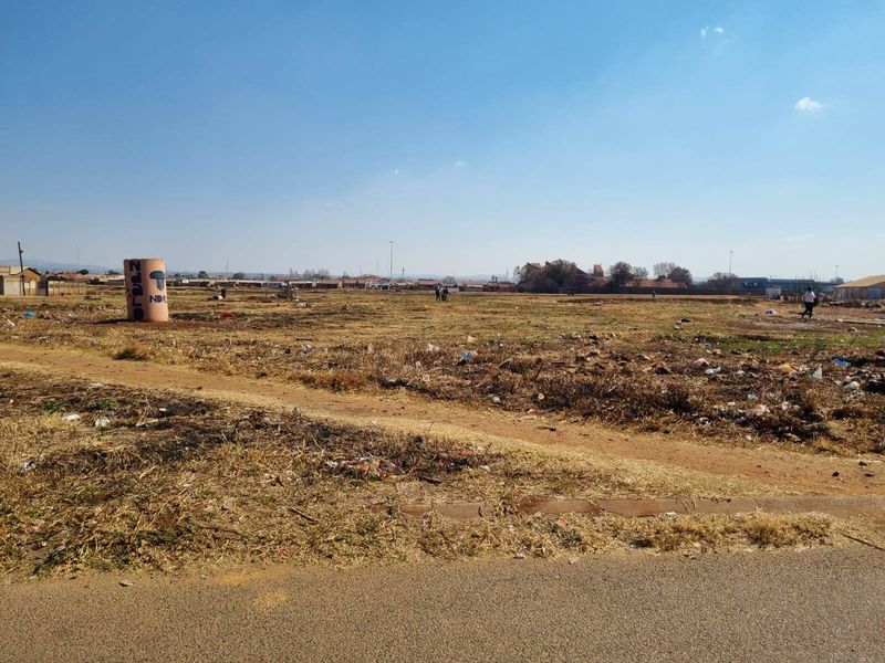 A wide-angle view of a vacant residential plot featuring dry, unmaintained ground and an unpaved access path.