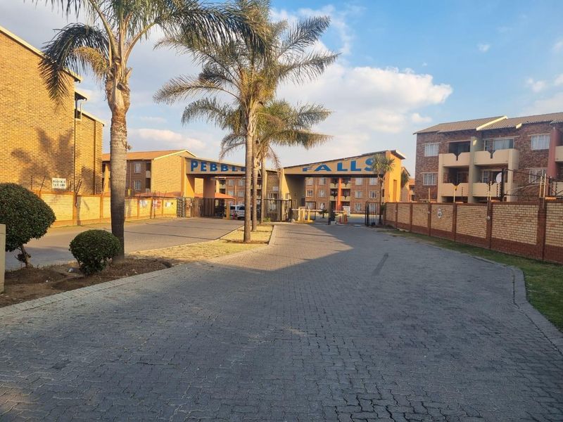 Entrance view of the Pebble Falls apartment complex featuring a paved driveway, secure gated access, and facebrick building exteriors.