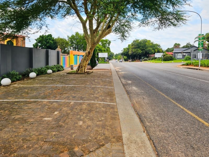 Street view showing a paved parking area adjacent to the road, featuring a boundary wall and mature tree.