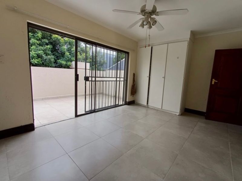 A spacious bedroom featuring tiled flooring, built-in cupboards, a ceiling fan, and direct access to an outdoor patio through a sliding glass door.