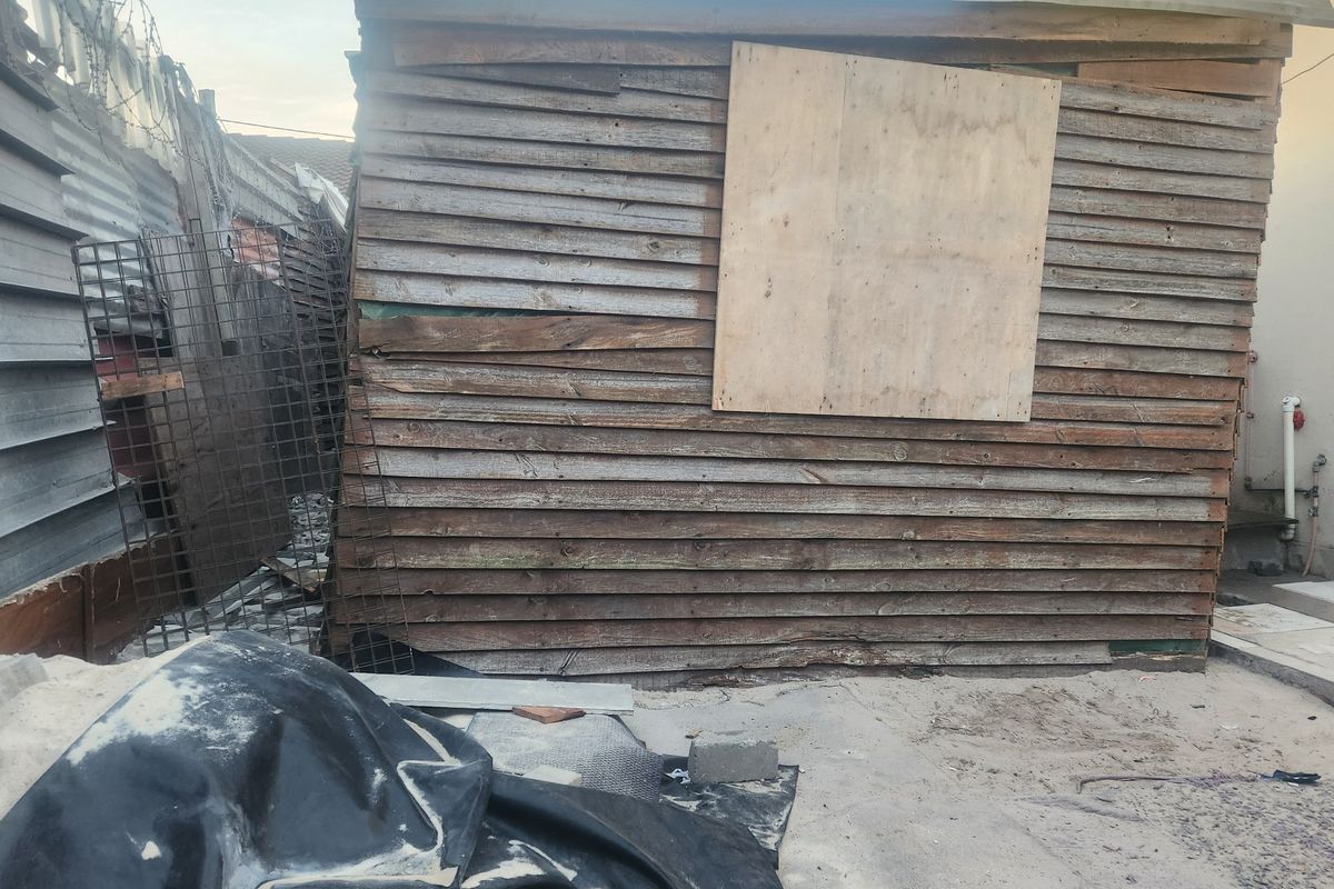 Rear exterior view showing a dilapidated timber structure with boarded window and surrounding metal fencing in poor condition.