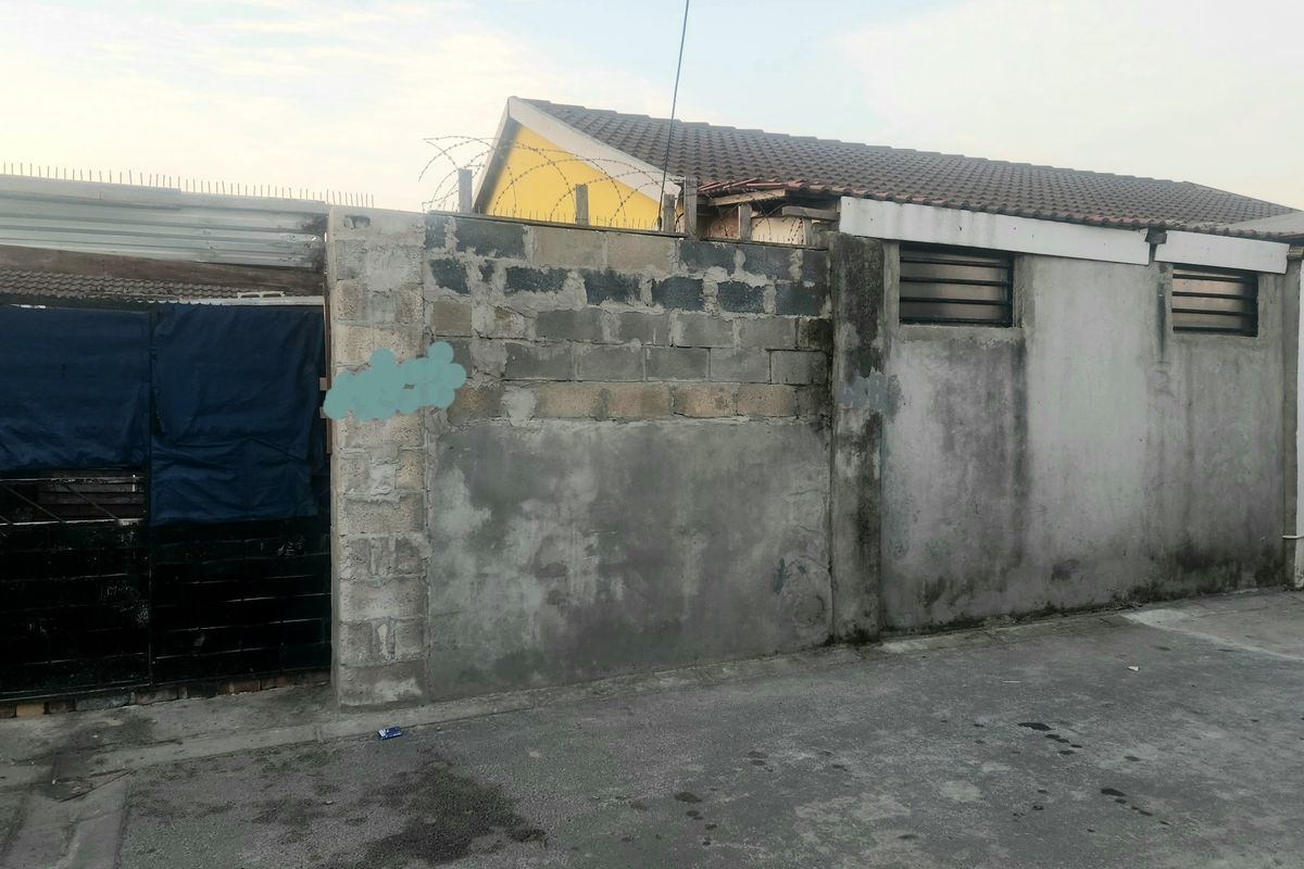 Exterior view of the property featuring a concrete boundary wall with razor wire, tiled roof, and louvered windows, showing signs of wear and unfinished surfaces.