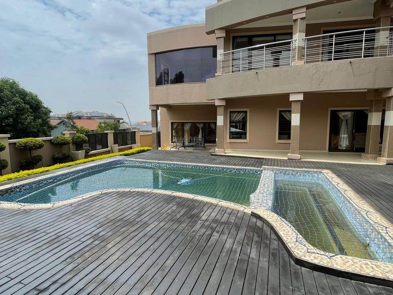 A spacious outdoor pool area featuring a multi-level swimming pool, extensive timber decking, and a two-story residential facade with a balcony.