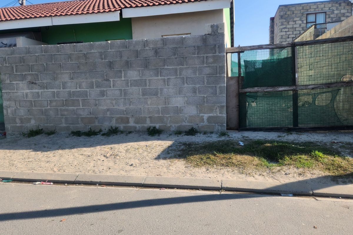 Street view of a residential property featuring a concrete block boundary wall, tiled roof, and a basic mesh gate entrance.