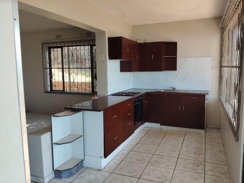 A functional kitchen featuring dark wood cabinetry, built-in oven and hob, and tiled flooring, opening into an adjacent living area.