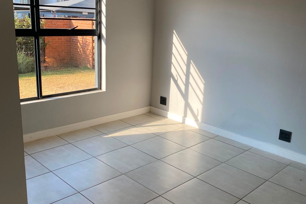 A bright, empty bedroom featuring neutral tiled flooring, clean grey walls, and a large window with black frames, offering a blank canvas for styling.