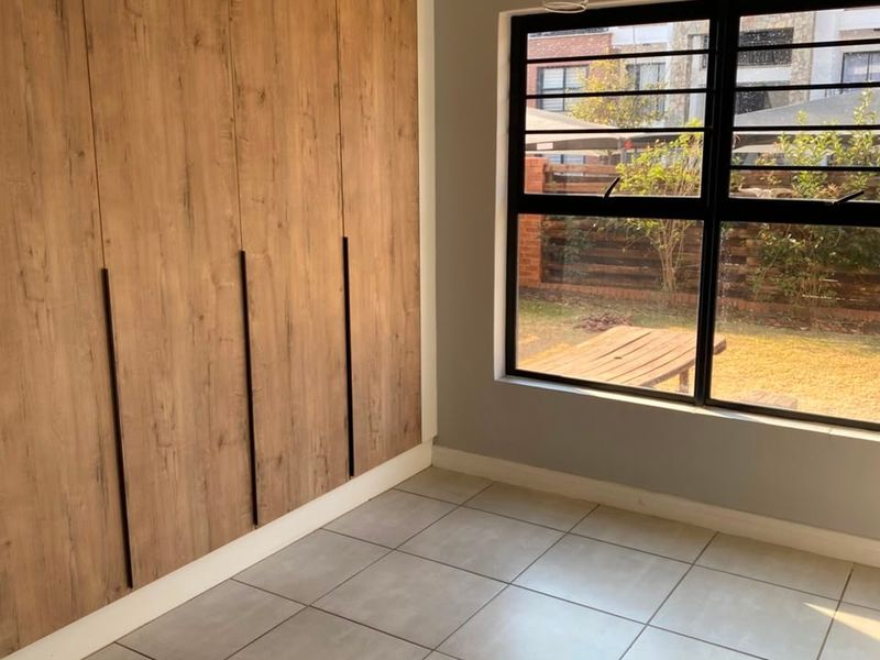 A bright, empty bedroom featuring modern wood-look built-in cupboards, neutral floor tiles, and a large window with black aluminium frames overlooking the garden.