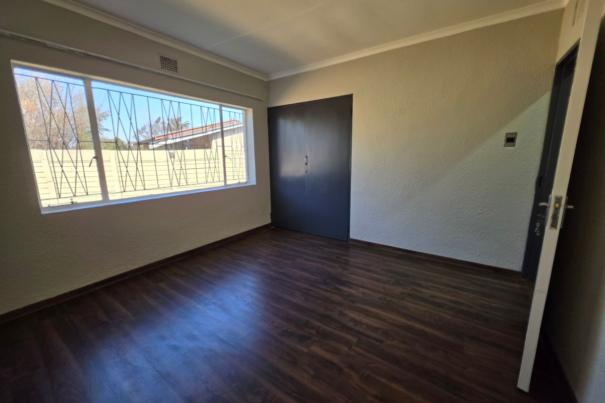 An empty bedroom featuring dark laminate flooring, a built-in cupboard, and large windows fitted with security bars.