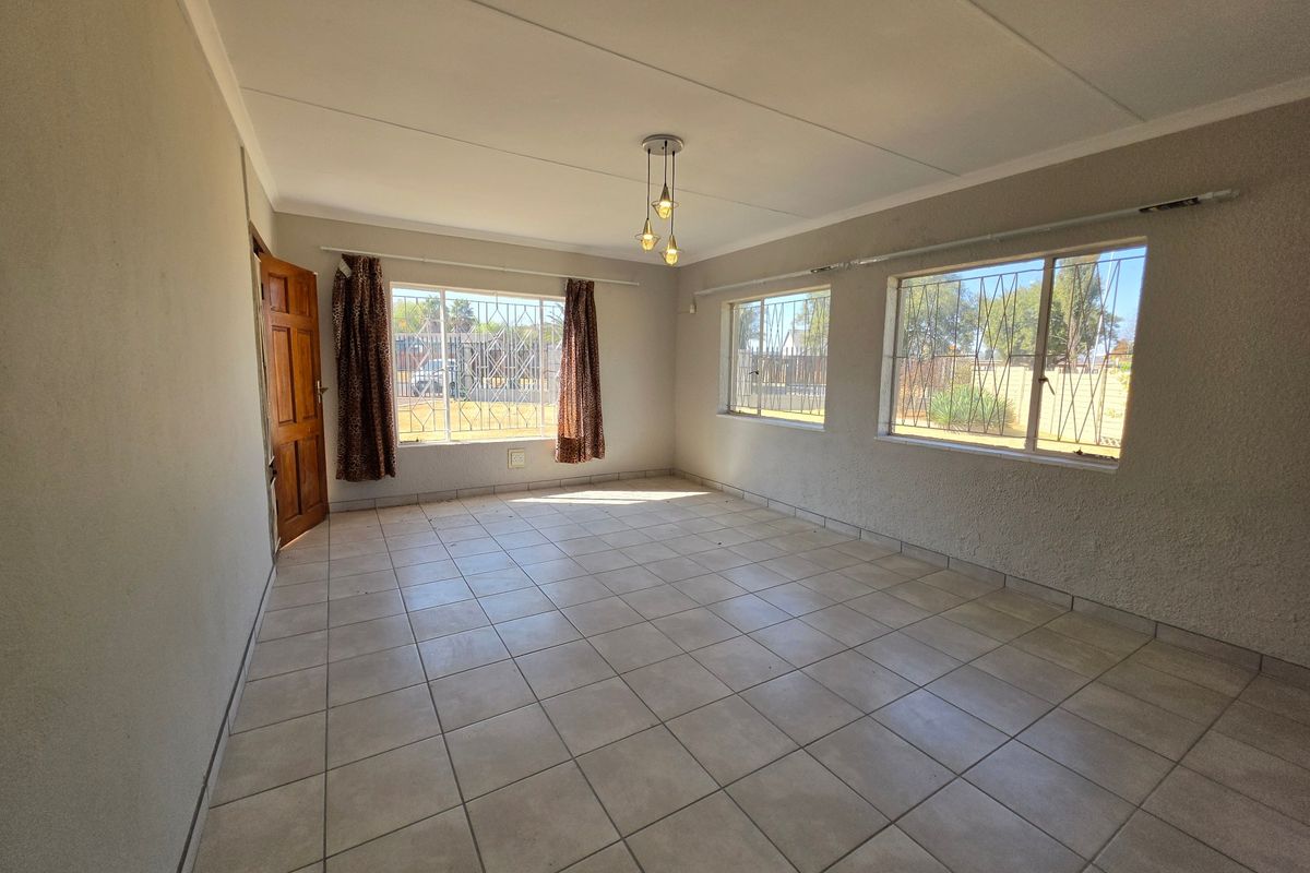 A spacious, empty living room featuring neutral tiled flooring, textured walls, and large windows fitted with security bars, offering a bright and versatile living space.