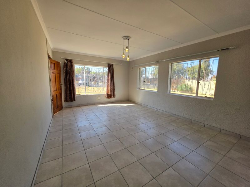 A spacious, empty living room featuring neutral tiled flooring, textured walls, and large windows fitted with security bars, offering a bright and versatile living space.