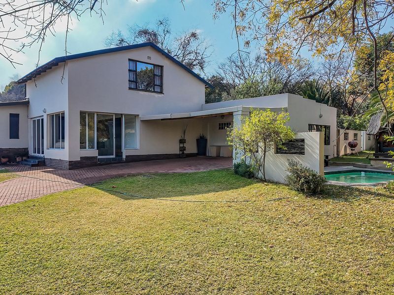Rear view of the property featuring a paved driveway, covered patio area, and a swimming pool set within a landscaped garden.