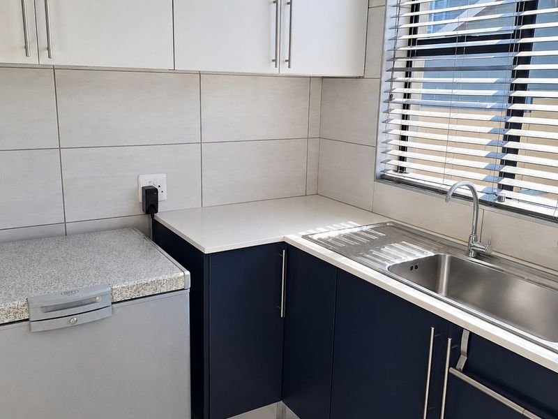 A modern, compact scullery featuring navy blue lower cabinetry, white upper cabinets, stone-look countertops, and a stainless steel sink with a tiled splashback.