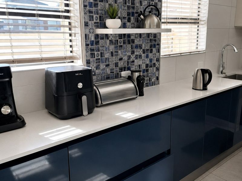 Modern kitchen featuring glossy navy cabinetry, white quartz countertops, and a decorative blue mosaic tile backsplash with floating shelving.
