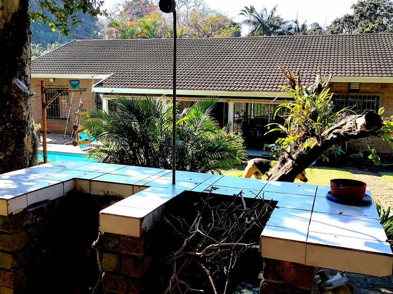 Rear garden view featuring a brick-built tiled braai area, swimming pool, and established landscaping with a view of the main house structure.