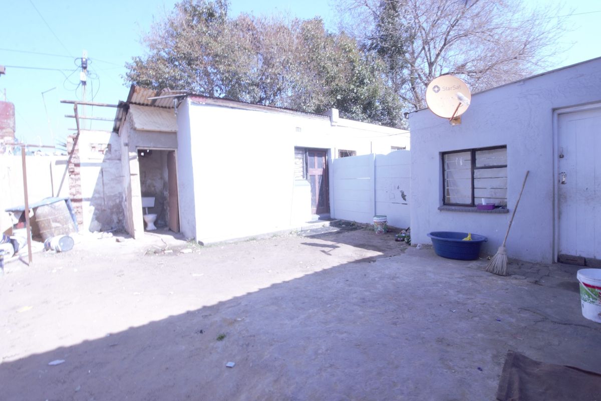 Rear exterior view of the property showing a basic yard area, external toilet structure, and plain masonry walls with visible signs of wear.