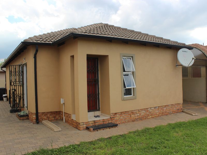 Rear view of a single-story house featuring a tiled roof, brick plinth, security gate, and paved walkway area.