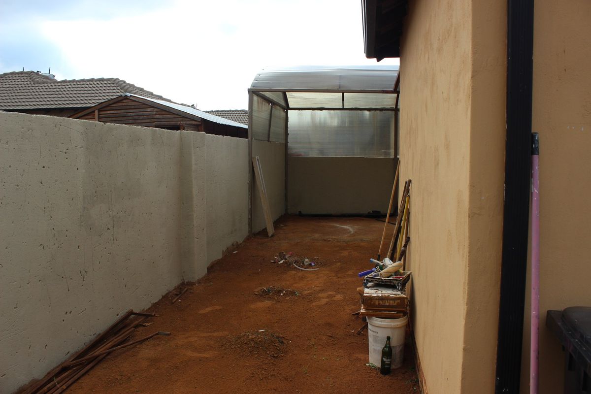 A narrow side or rear yard area featuring a boundary wall and a simple polycarbonate roof structure over an unfinished dirt ground surface.