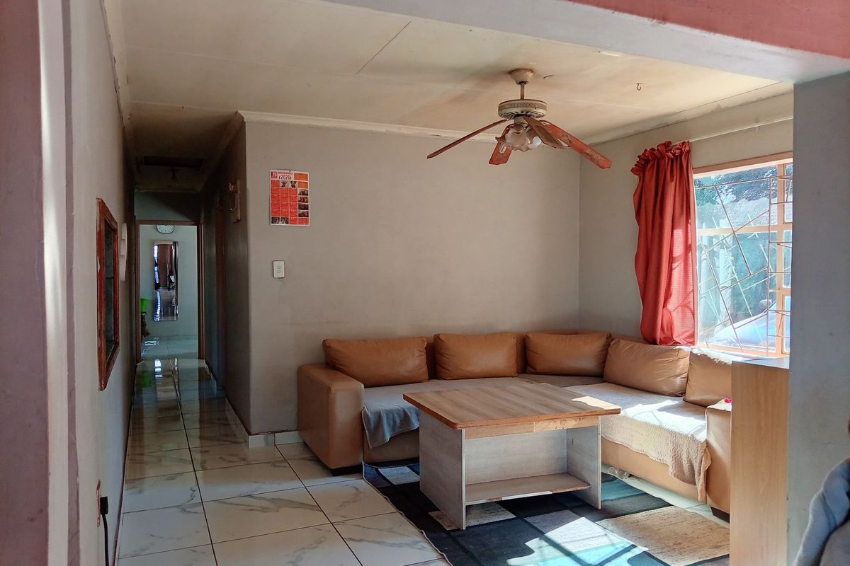 A living room featuring tiled flooring, a ceiling fan, and security bars on the window, showing signs of wear on the ceiling.