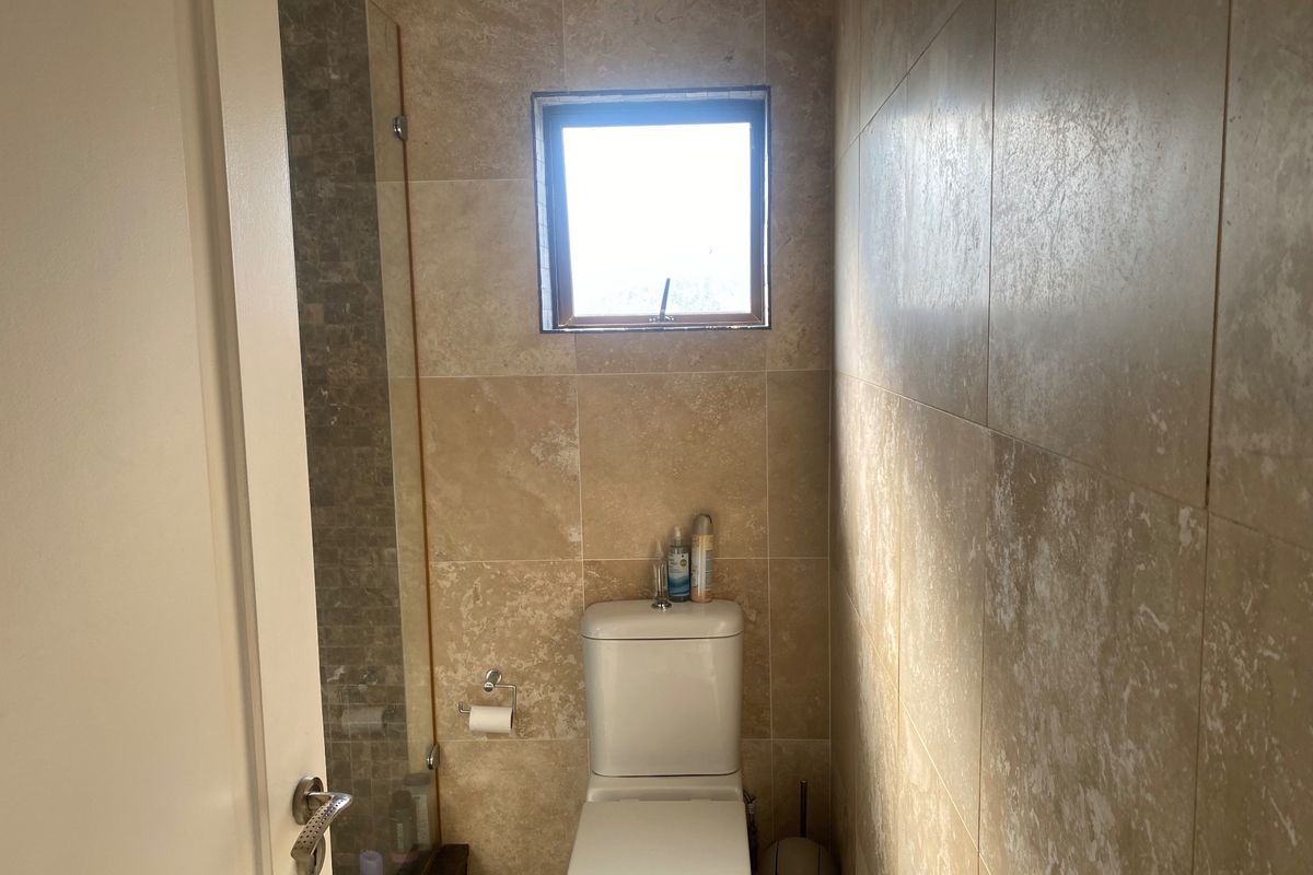 A compact bathroom featuring floor-to-ceiling travertine wall tiles, a modern toilet, and a glass-enclosed shower area with a small window for natural light.