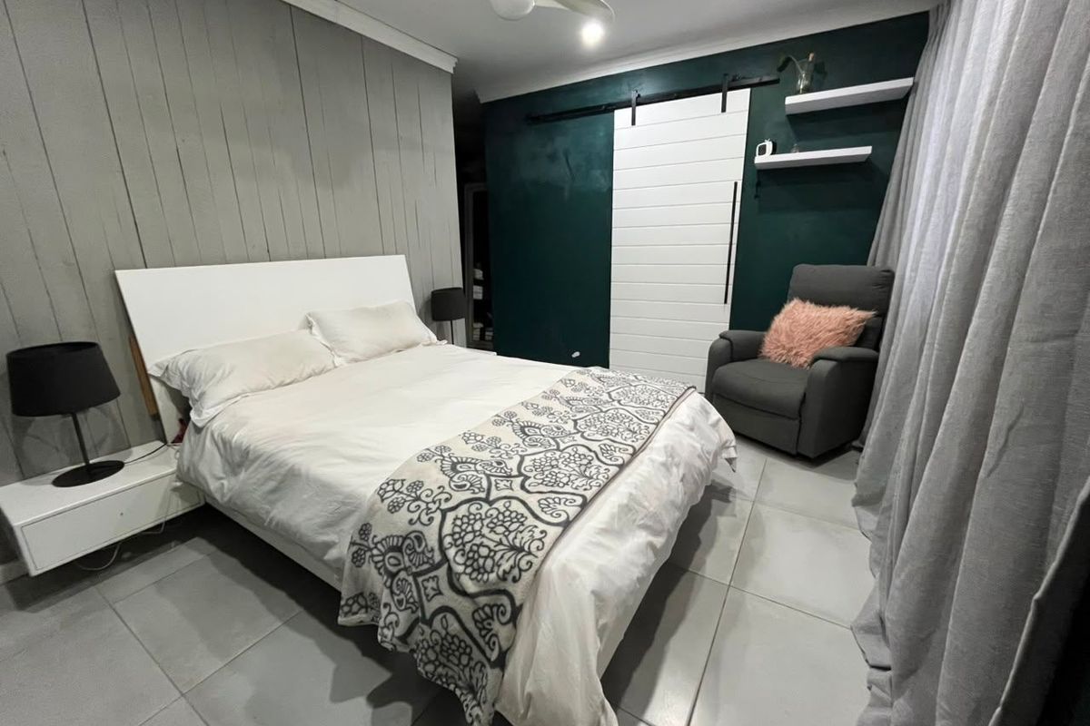 A well-maintained bedroom featuring a modern sliding barn door, ceiling fan, floating wall shelves, and neutral tiled flooring.