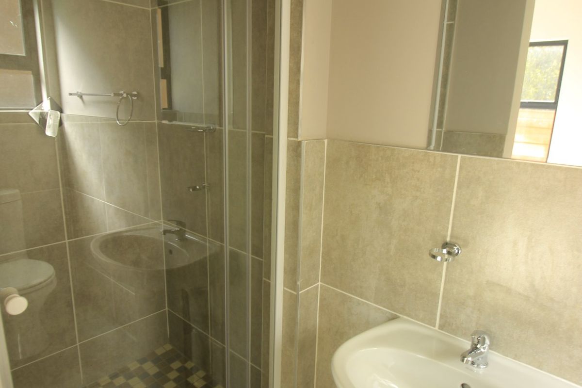 A clean, modern bathroom featuring a pedestal basin, wall-mounted mirror, and a glass-enclosed shower with neutral-toned wall and floor tiling.