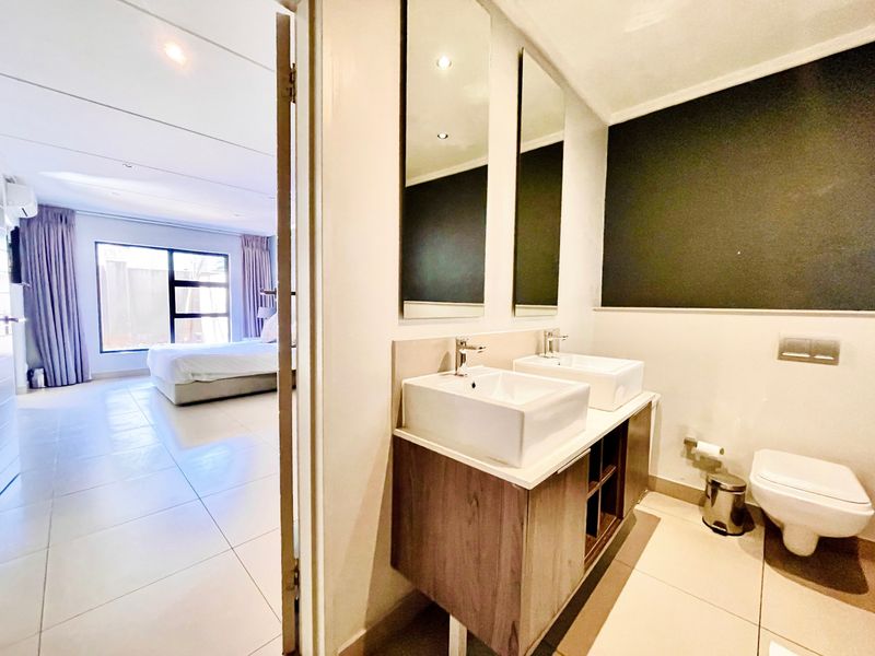 Modern bathroom featuring a double vanity with vessel sinks, wall-mounted toilet, and large mirrors, adjacent to a bright bedroom.