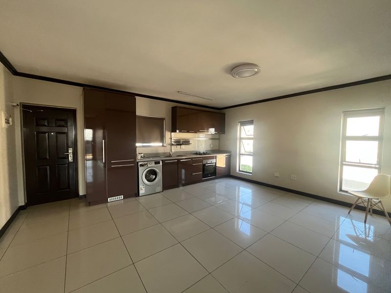 Open-plan living area featuring a modern kitchen with glossy brown cabinetry, built-in oven, hob, and integrated laundry facilities, finished with neutral floor tiles.