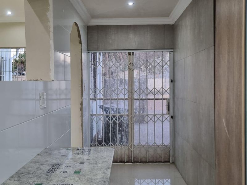 An empty kitchen area featuring stone countertops, tiled walls, and a security gate leading to the exterior, currently requiring finishing touches and maintenance.